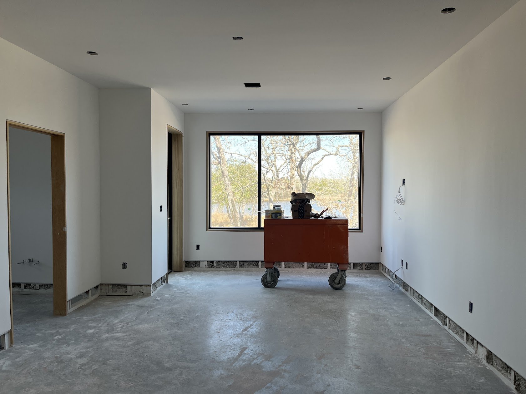 A bedroom under construction.  There is a large window overlooking a lake and a toolbox in the center of the room.